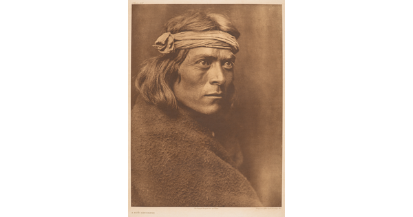 A sepia-toned portrait photograph of an Indigenous man wrapped in a blanket and wearing a cloth wrap on his head.