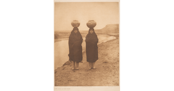 A sepia-toned photograph of two Indigenous women wrapped in blankets standing on a riverbed with water jugs balanced on their heads.