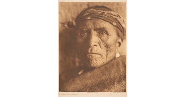 A sepia-toned portrait photograph of an older Indigenous person wearing a headband, and a solid-colored cloth wrapped around their shoulders and under their face.