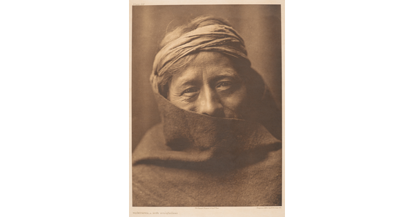 A sepia-toned portrait photograph of an older Indigenous person wearing headband; their face partially covered by what looks like a blanket.