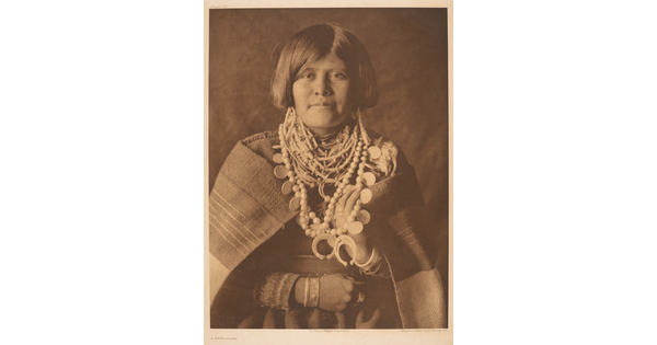 A sepia-toned portrait photograph of a young Indigenous person with short, bobbed hair, wearing many beaded necklaces, and a blanket wrapped around her shoulders.