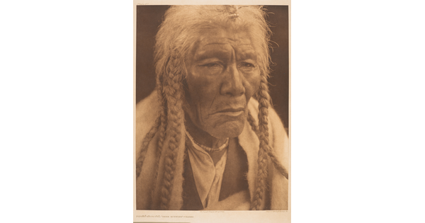 A sepia-toned portrait photograph of an elderly Native American man with long, gray hair in several braids gazing out.