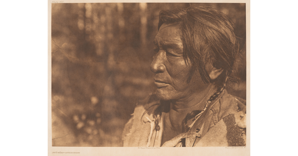 A sepia-toned photograph of a middle-aged Native American man's face in profile; short hair in two braids.