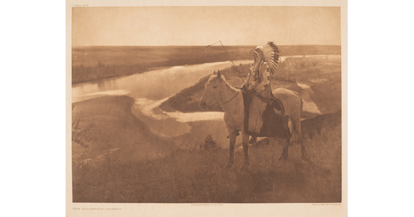 A sepia-toned photograph of a Native American man, in profile and wearing a full feathered headdress, on a horse looking off to a river in the background.