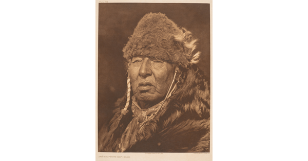 A sepia-toned portrait photograph of an older Indigenous person wearing fur clothing and a fur hat.
