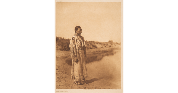 A sepia-toned photograph of an Indigenous woman with her hair pulled back into a braid, wearing fringed clothing that looks to be made of animal skins, standing on a shoreline.