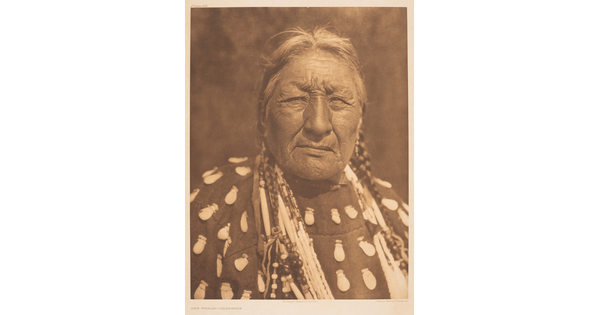 A sepia-toned portrait photograph of an older Indigenous woman wearing a braids, elk-tooth shirt, and multiple necklaces.