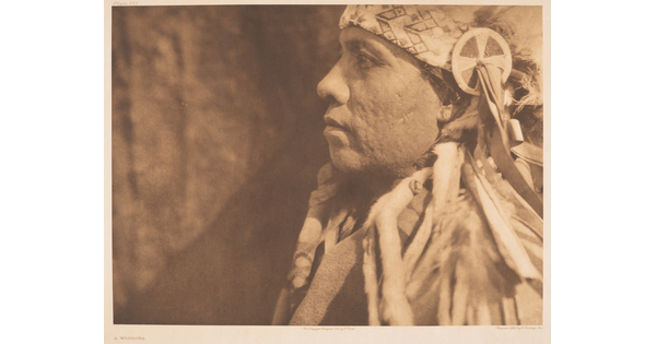 A sepia-toned, close-up, photograph of an Indigenous man in profile wearing a traditional headdress.