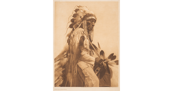 A sepia-toned portrait photograph of an older Indigenous man wearing a feathered headdress and cultural clothing.