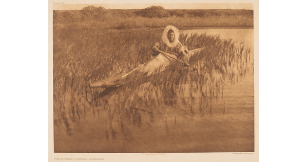 A sepia-toned photograph of an Indigenous person with a fur-lined hood pulled up around their head in a kayak among reeds on the water.