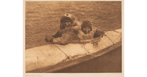 A sepia-toned photograph of two Indigenous people bundled up sitting in a kayak on the water.