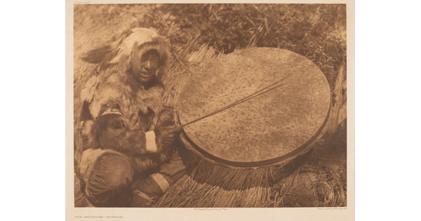 A sepia-toned photograph of an Indigenous person wearing furs squatting next to a large drum they are hitting with a stick.