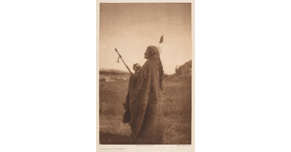 A sepia-toned photograph of an Indigenous adult standing on a grassy field wrapped in animal skins.
