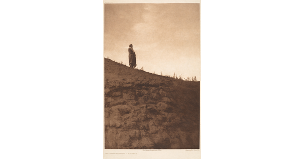 A sepia-toned photograph of an Indigenous person standing high on a hill with a blanket around their shoulders.