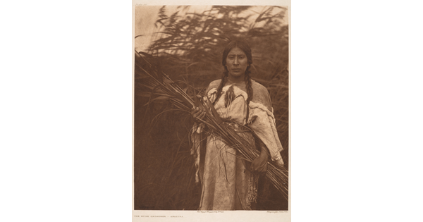 A sepia-toned photograph of an Indigenous woman, hair in braids and wearing a traditional dress, holding a large bundle of grasses in her hands.