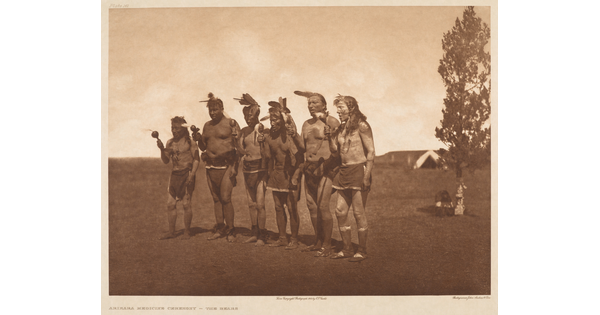 A sepia-toned photograph of six Native American men standing shoulder-to-shoulder, all holding something in their right hands.