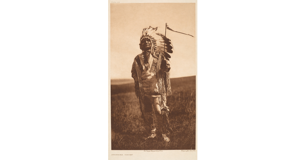 A sepia-toned photograph of an Indigenous man standing in a grassy field wearing a feathered headdress and ornately decorated traditional clothing.