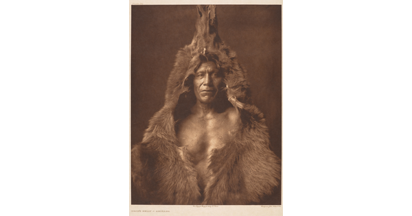 A sepia-toned portrait photograph of a Native American man wearing a bear skin over his head and shoulders.