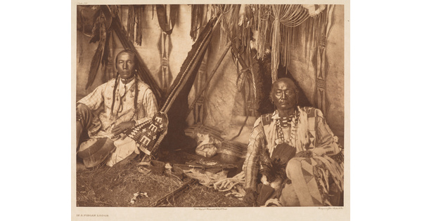 A sepia-toned photograph of two Indigenous adults sitting inside a Native American lodge wearing cultural dress.