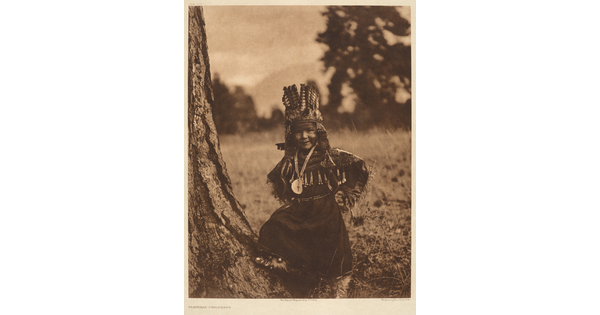 A sepia-toned portrait photograph of a Native American child in traditional dress and headdress, standing next to a tree with hands on hips.