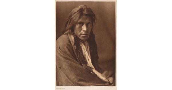 A sepia-toned portrait photograph of an Indigenous man with a blanket around his shoulders leaning toward the camera.