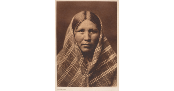 A sepia-toned portrait photograph of an American Indian woman, a plaid shawl over most of her head and shoulders.