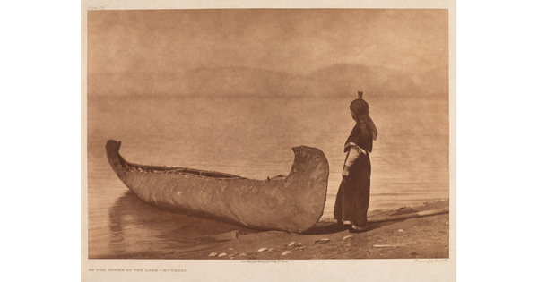 A sepia-toned photograph of a canoe on the edge of the shore and an Indigenous person standing beside it.