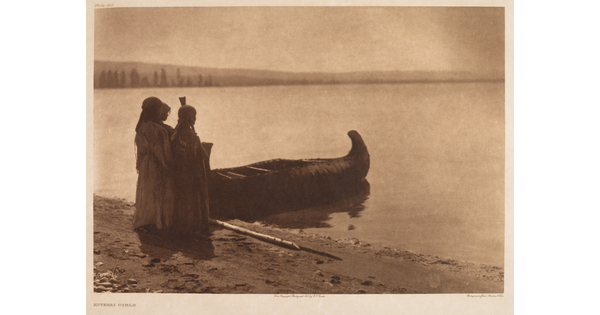 A sepia-toned photograph of three Indigenous people standing on a shore next to a canoe on calm water.
