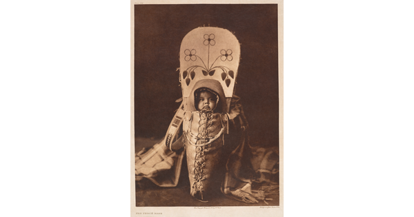 A sepia-toned portrait photograph of an Indigenous baby in a large cradleboard.