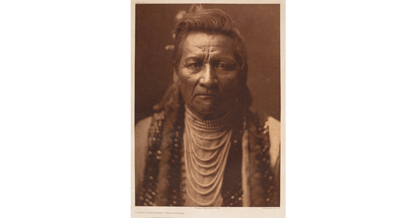 A sepia-toned portrait photograph of a Native American man from the chest up in traditional dress, looking at the viewer.