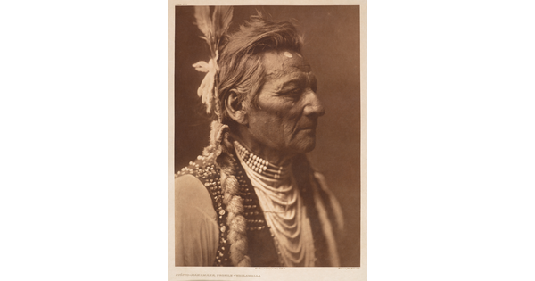 A sepia-toned portrait photograph of a Native American man in profile from the shoulders up wearing traditional dress.