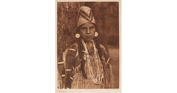 A sepia-toned portrait photograph of a Native American girl with braids, wearing a tall decorative hat and traditional clothing.
