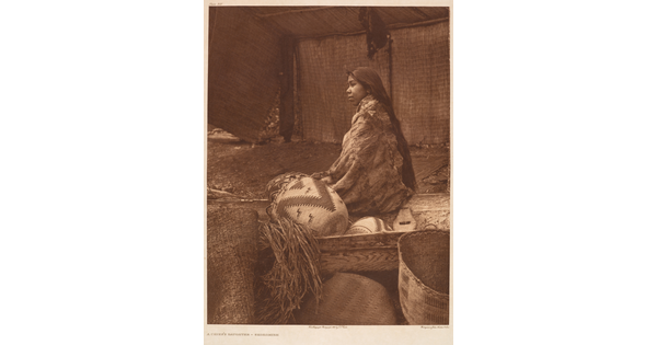 A sepia-toned photograph of a Native American woman wrapped in a blanket, sitting with back to the viewer next to a wood crate containing woven baskets.