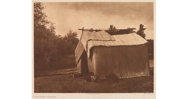 A sepia-toned photograph of a wood structure covered with woven straw mats and a few pots at the entrance.