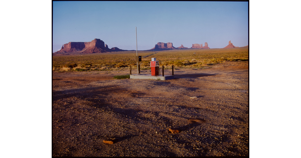 A color photograph of a lone gas pump on a concrete pad in a desert landscape with mesas and buttes in the background.