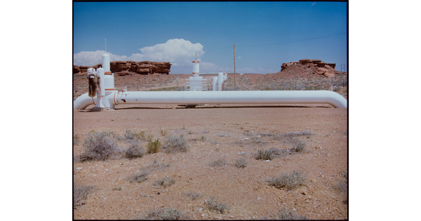 A color photograph of a white pipes and other machinery in a desert landscape with red rock formations in the background.