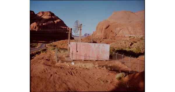 A color photograph of a small rectangular structure with a satellite behind a chain-link fence in the middle of a desert with many red rock formations.