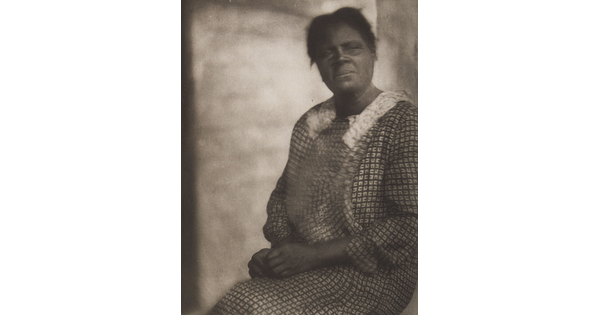 A black-and-white, unfocused photograph of a seated Black woman in a patterned dress, gazing out.