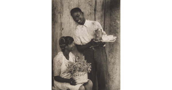 A black-and-white photograph of a young Black woman holding a basket of flowers on her lap and a young Black man standing next to her with a coffee service, both smiling.