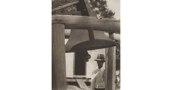 A black-and-white photograph of a Black man in a hat standing to the side of a wooden structure that holds a large bell.