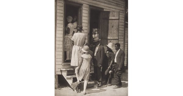 A black-and-white photograph of a group of Black people dressed nicely going up steps into a clapboard building.