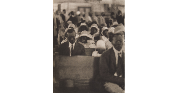 A soft-focus, black-and-white photograph of Black congregants seated in crowded church pews and standing in the back.