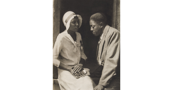 A black-and-white photograph of a young Black couple dressed nicely and seated in a doorway.