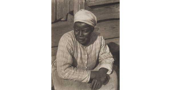 A black-and-white photograph of a Black woman, head covered in a white kerchief, wearing a plaid dress seated on a wood stoop with her arms crossed on her lap.