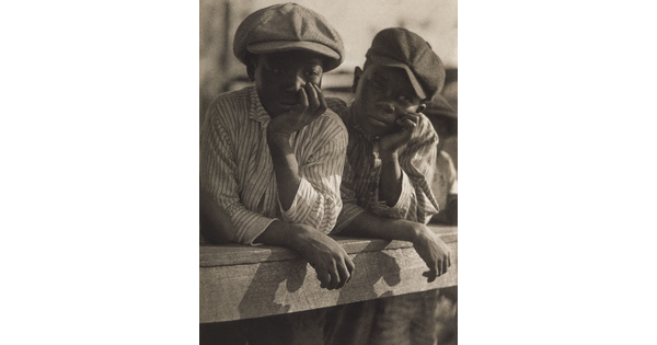 A black-and-white photograph of two young Black boys, both wearing newsboy caps and striped shirts, and both leaning on a wood fence, chins resting in hands.