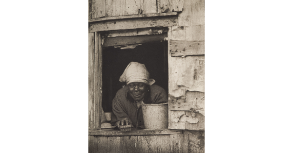 A black-and-white photograph of a Black woman wearing a bandana over her hair, holding a scrub brush and leaning over a pail from the window of a wood building.