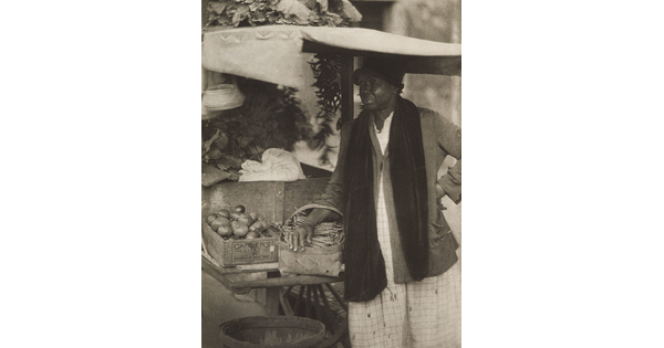 A black-and-white photograph of a Black woman standing with a basket on her arm next to a cart of crated vegetables.