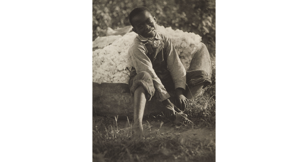 A black-and-white photograph of a smiling young Black boy sitting on a bag of cotton.