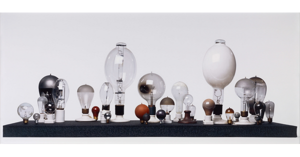 A color photograph of lightbulbs of many shapes and sizes, arranged with their socket sides down on a black platform in front of a white background.