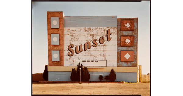 A color photograph of a weathered marquee sign for a drive-in movie theater with the word &quot;Sunset&quot; in script.